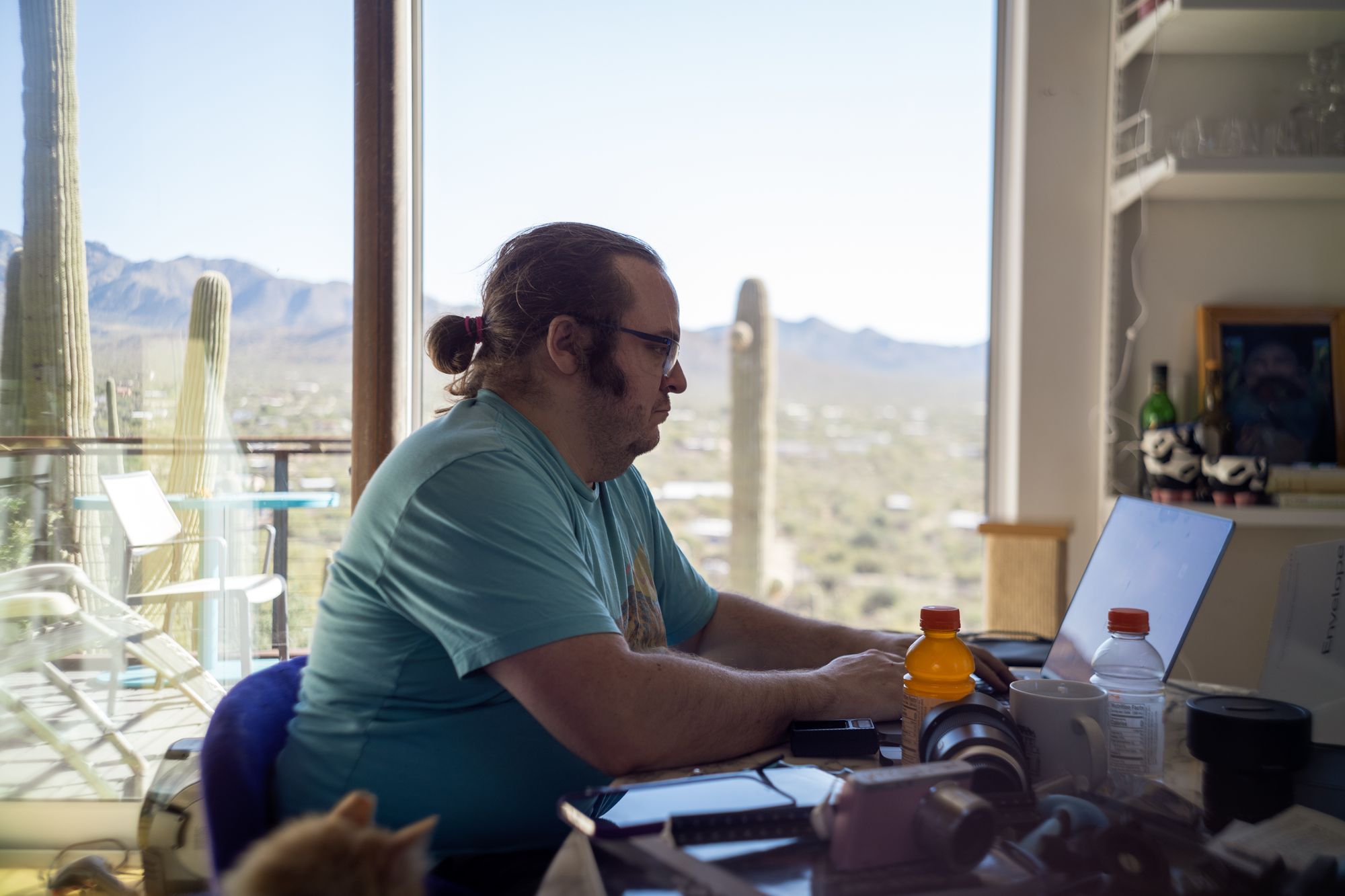 vintage-y looking photo of a man at a dining table piled with stuff, looking at a laptop; clearly in focus but soft in a pleasing way